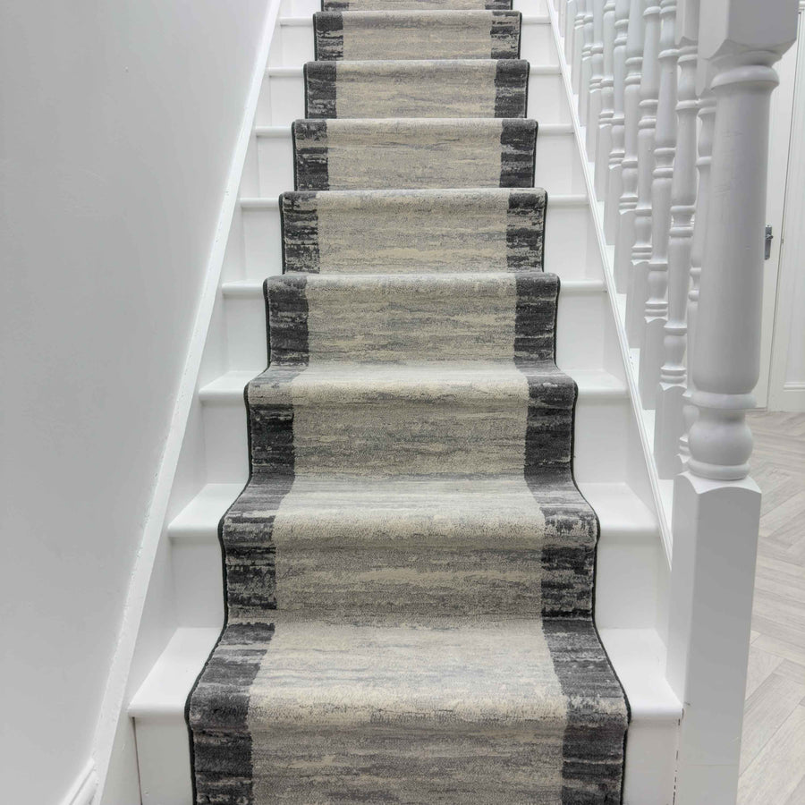 A staircase with white steps and a hallway featuring the Eclipse 79138-7696 Runner Rug, which has a gradient pattern in gray and beige tones, flanked on the right by white banisters.