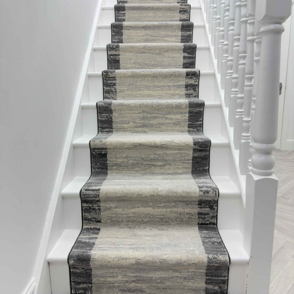 A staircase with white steps and a hallway featuring the Eclipse 79138-7696 Runner Rug, which has a gradient pattern in gray and beige tones, flanked on the right by white banisters.