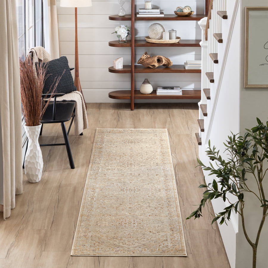 A modern, neutral-toned hallway with a light beige runner rug on wood floors, a black chair with a blanket, potted plants, and a curved wooden bookshelf displaying decorative items. Stairs and framed art are on the right.