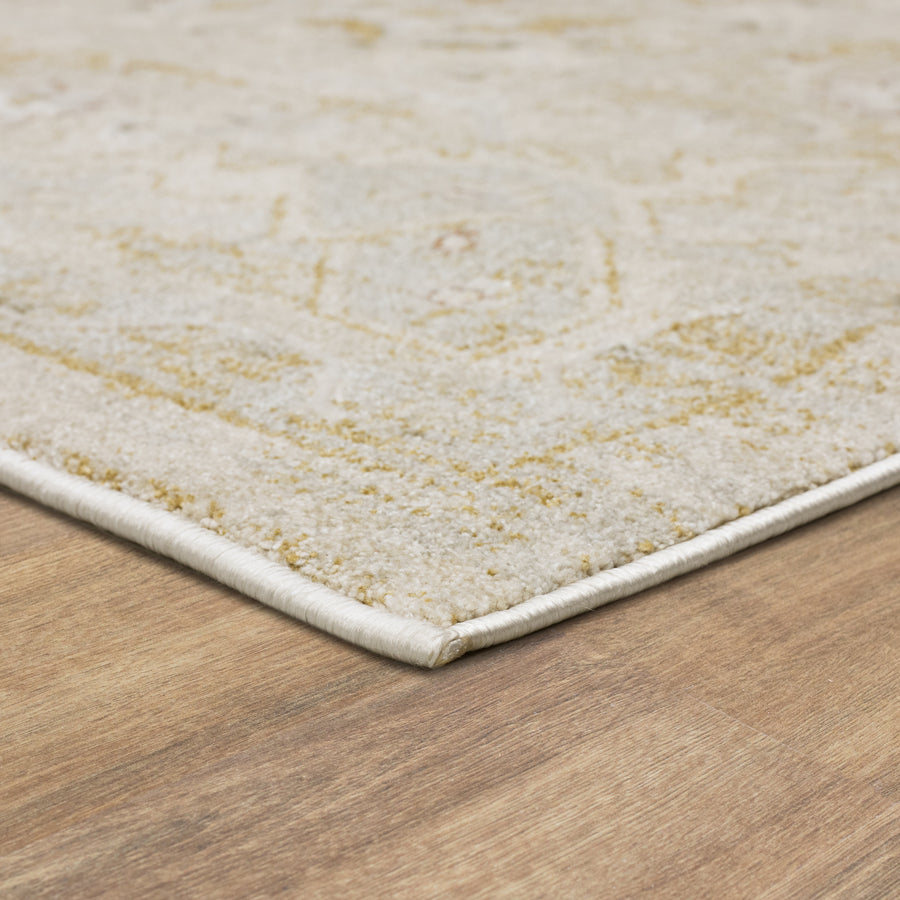 Close-up of the corner of a light-colored, patterned area rug with beige and yellow tones, placed on a wooden floor. The rug’s edge is neatly finished and slightly lifts off the surface.