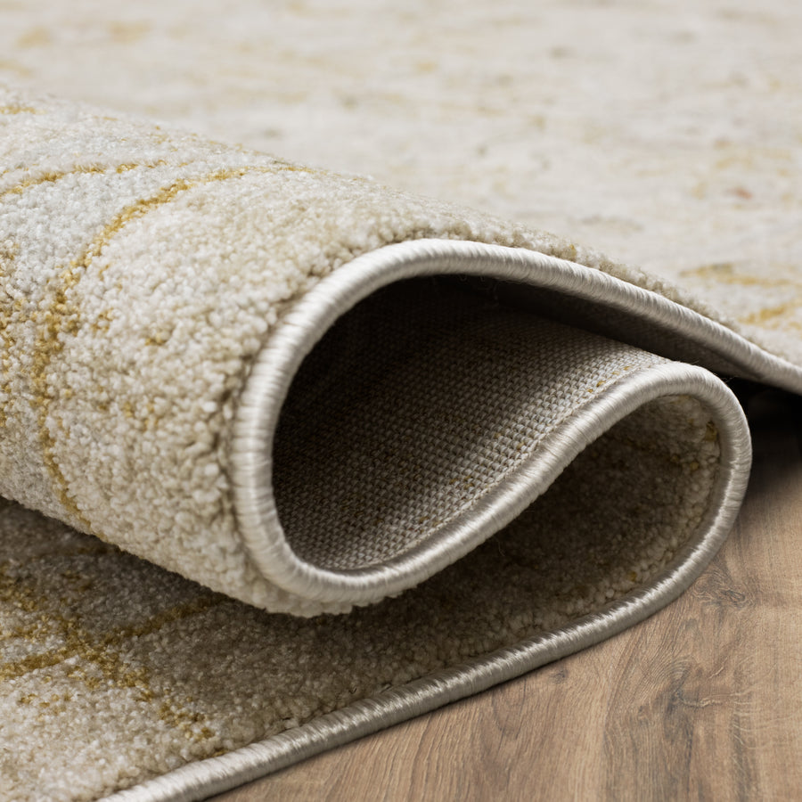 A close-up of a beige and cream patterned rug with its corner partially folded over on a wooden floor, showing the texture and underside of the rug.