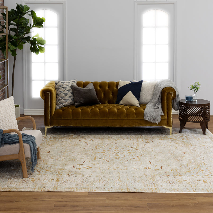 A living room with a mustard yellow velvet tufted sofa, patterned pillows, a gray throw, a round wooden side table, an armchair, a large light-patterned rug, a tall potted plant, and two arched windows with sheer curtains.
