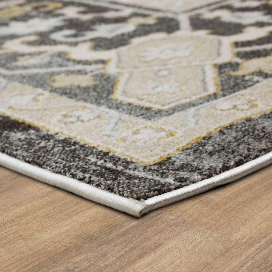 A close-up of the corner of a patterned area rug with a beige, gray, and black design, placed on a wooden floor. The rug has a white edge binding and a soft, textured surface.
