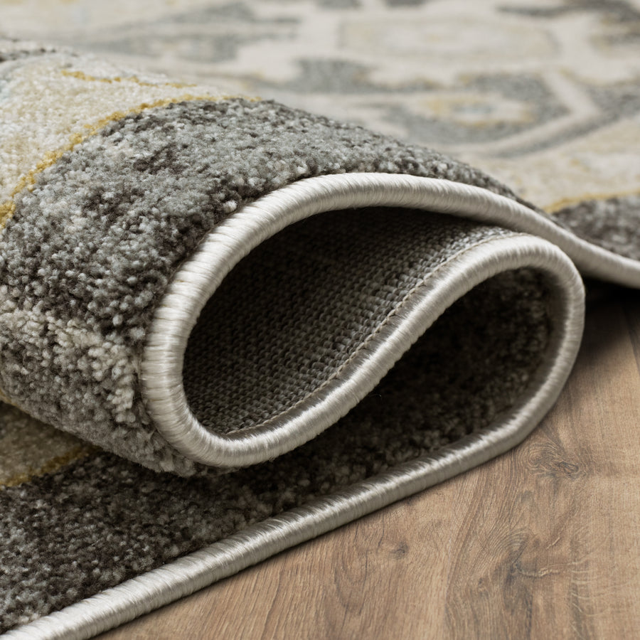 A close-up view of the edge of a rolled-up rug with a textured, patterned surface in shades of gray and beige, resting on a wooden floor.