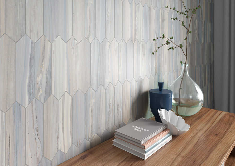 A wooden table with stacked books, a white decorative shell, a blue jar, and a glass vase with branches sits in front of a wall with hexagonal tiles in muted blue and beige tones.