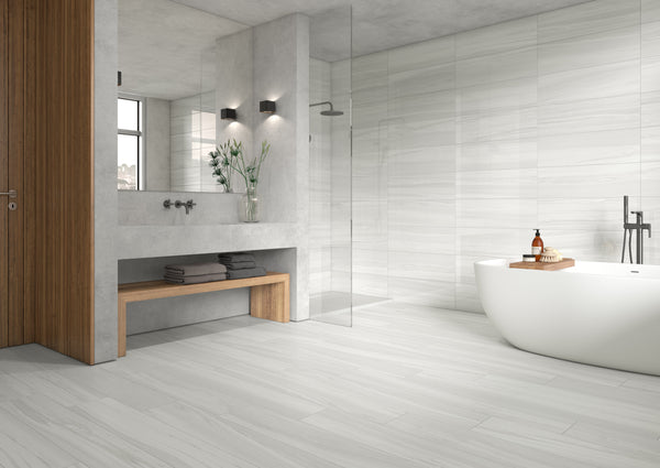 Modern bathroom with light gray marble tiles, a freestanding white bathtub, glass shower, wooden door, built-in concrete sink with mirror, and a wooden bench with towels underneath. Minimalist decor with a vase of flowers.