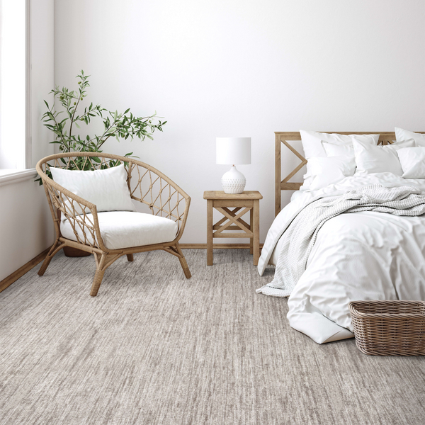 A cozy bedroom with a rattan chair, white cushions, a bedside table with a lamp, a neatly made bed with white bedding, a woven basket, and a potted plant by a window. The decor is light and neutral.