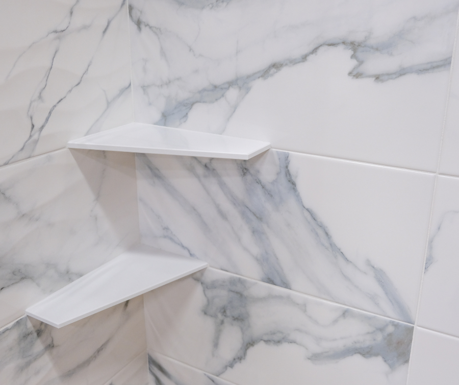 Two white corner shelves are installed on a wall covered with white marble-patterned tiles. The shelves are placed one above the other in a shower or bathroom setting.
