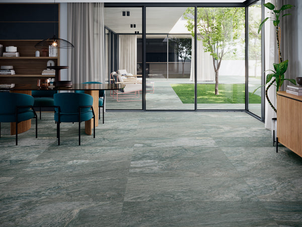Modern living and dining area with greenish marble flooring, blue chairs, wooden furniture, and large glass doors opening to a patio with trees and outdoor seating. Ample natural light fills the space.