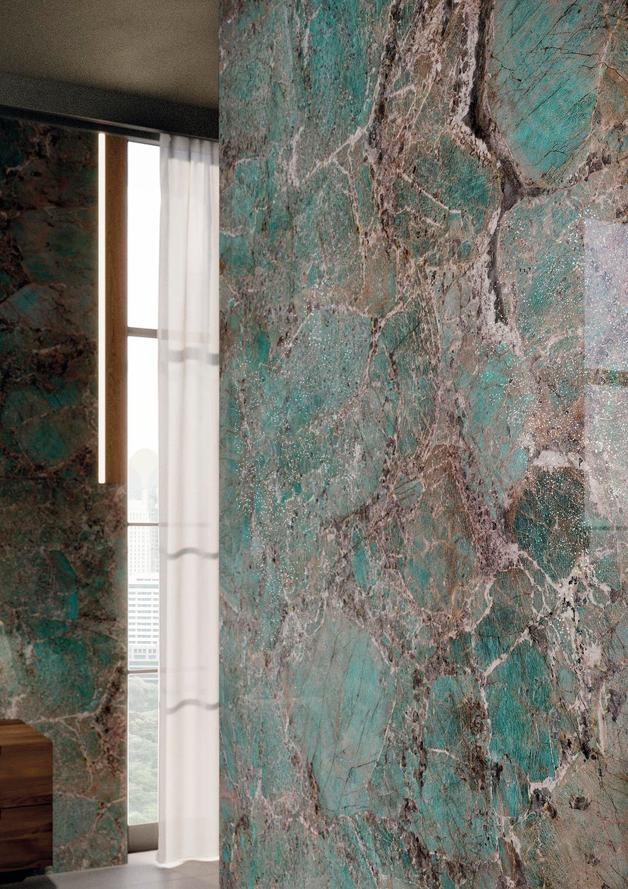 A close-up of a wall covered with teal and brown natural stone tiles, featuring large, irregular patterns. In the background, sheer white curtains partially cover a tall window with a cityscape view.