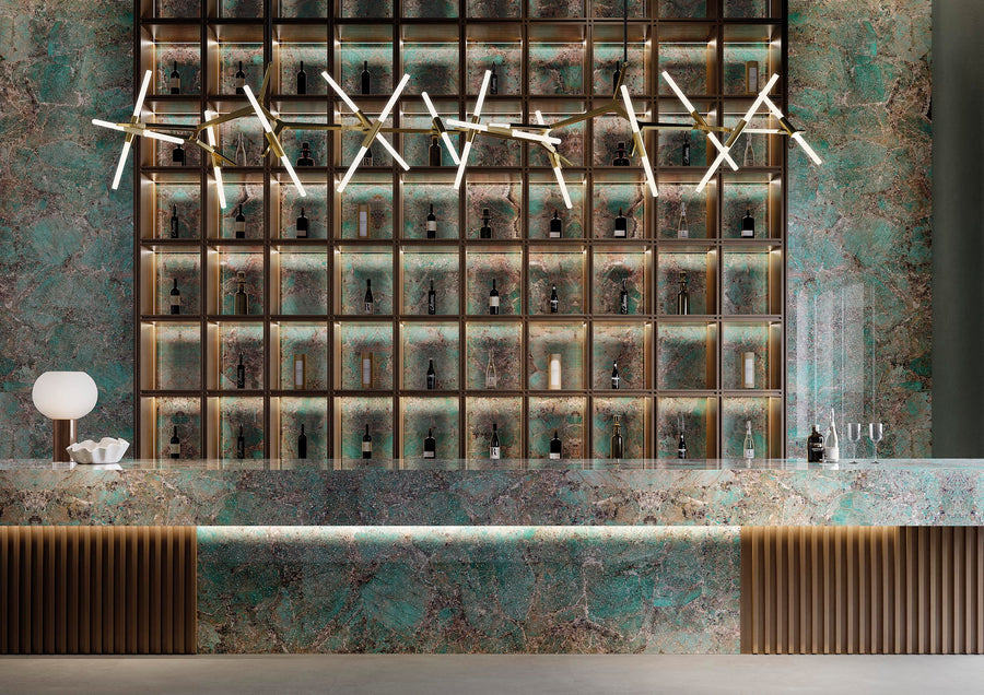 A modern bar with a marble-patterned green and brown counter, illuminated shelves displaying bottles, a geometric ceiling light fixture, a white lamp, bowls, and empty glasses on the counter.