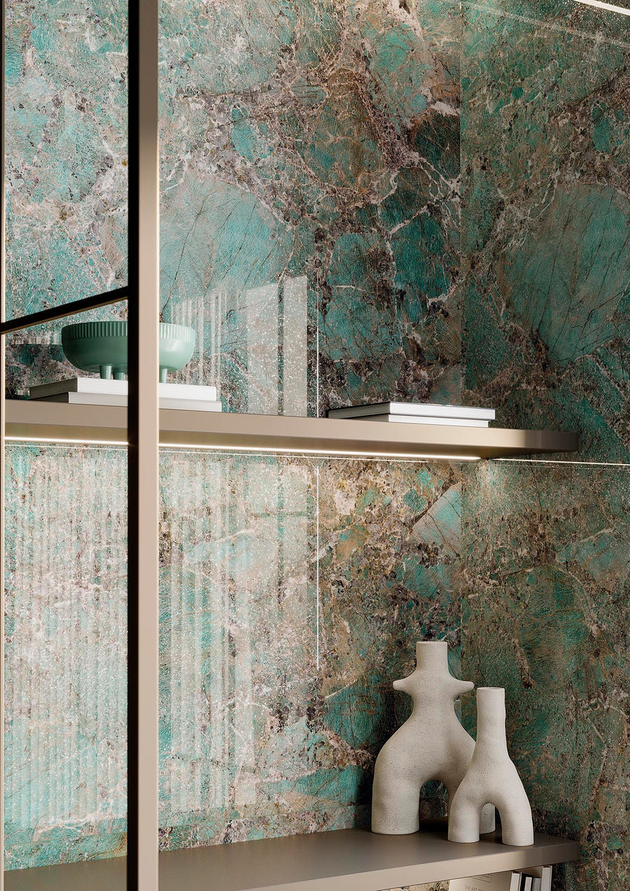 A modern shelf with white books, a green bowl, and two off-white abstract vases is set against a decorative green and brown marbled stone wall.