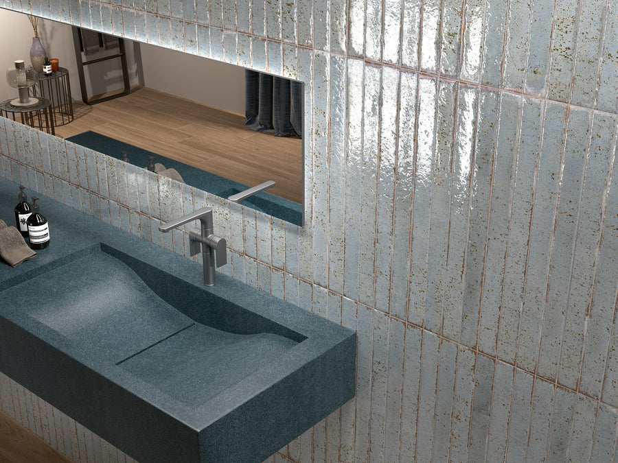 Modern bathroom with a long blue textured sink, a sleek metal faucet, and glossy light blue tiles on the wall. A large horizontal mirror reflects part of the room and wood flooring. Toiletries sit by the sink.