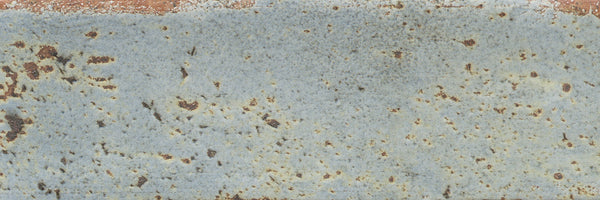 Close-up of Emerge 3x8 Porcelain Tile in Aurora color, showing a weathered look with softly worn edges, patches of brown and blue-gray tones, and scattered spots for natural variation.