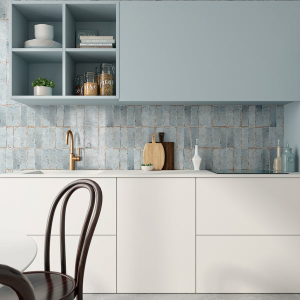 Modern kitchen with white cabinets, light blue upper cabinets, and a textured blue brick backsplash. There are open shelves with jars and a plant, a gold faucet, cutting boards, and a dark wooden chair.