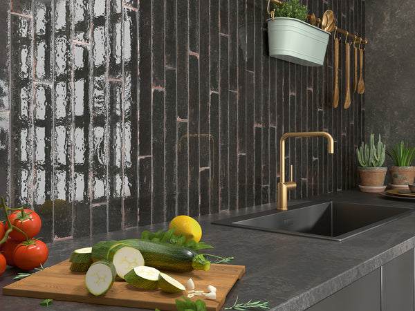 A modern kitchen countertop with a black sink and gold faucet, fresh vegetables on a cutting board, potted plants, and utensils hanging on a dark tiled backsplash.