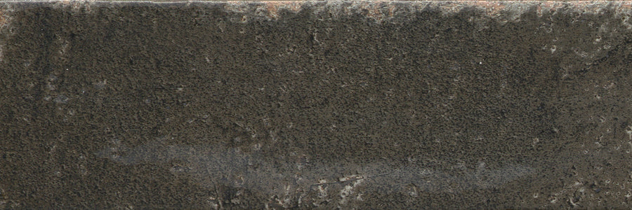 A close-up of Emerge 3x8 Porcelain Tile in Eclipse reveals a dark, weathered surface with natural variation, rough texture, softly worn edges, and subtle patches of lighter gray and brown accents.