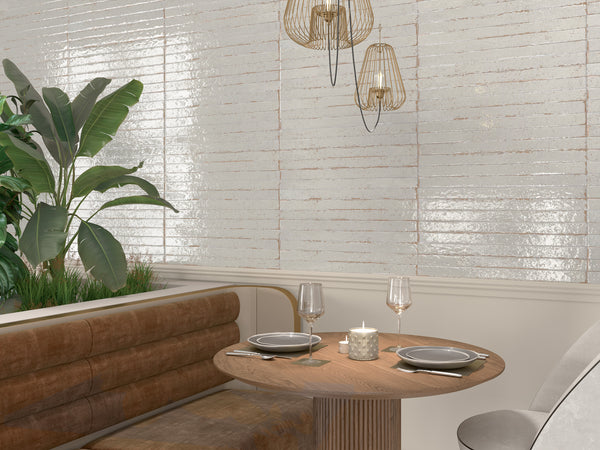 A cozy restaurant seating area with a round wooden table set for two, a lit candle, brown upholstered bench, decorative pendant lights, a gray chair, and green plants against a textured white tiled wall.
