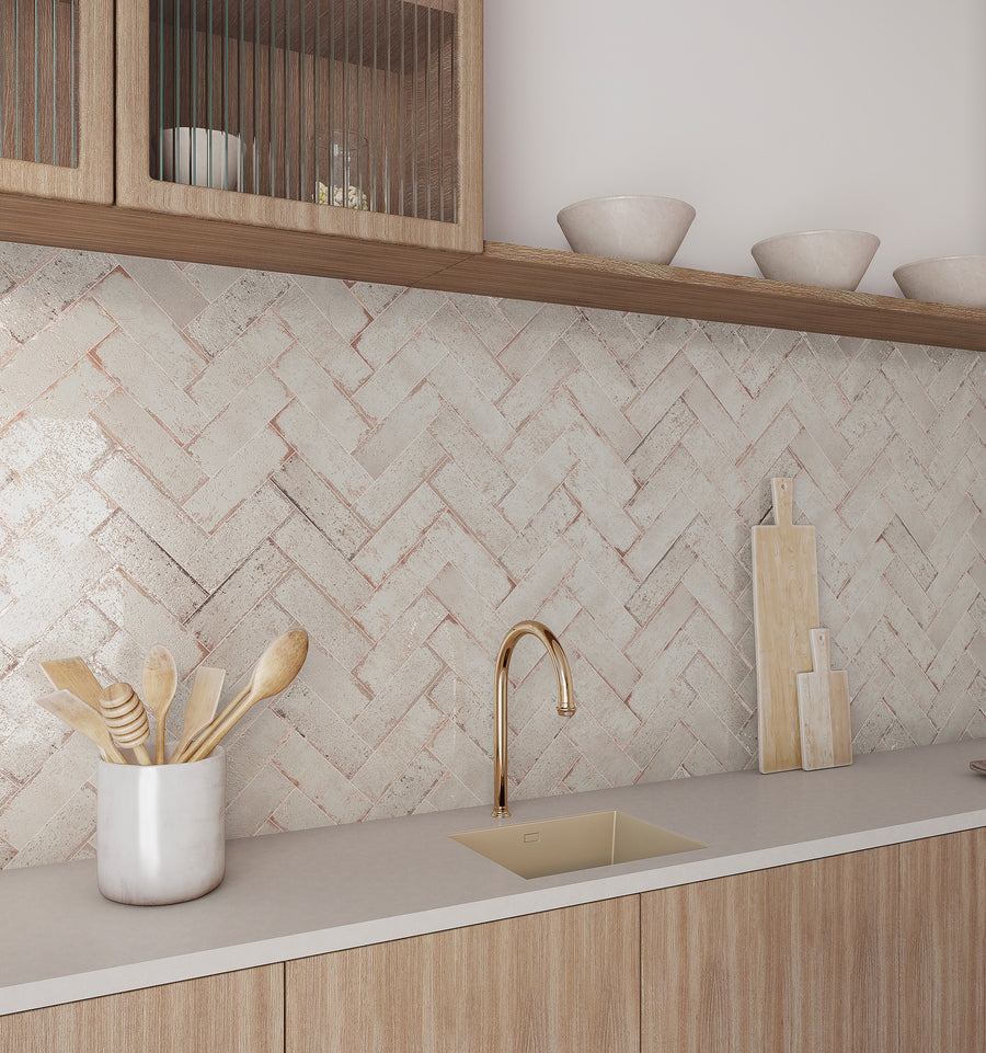 Modern kitchen with light wood cabinets, beige countertop, gold faucet, and herringbone-patterned backsplash tiles. Utensils are in a metal holder, with cutting boards and ceramic bowls on the counter and upper shelves.