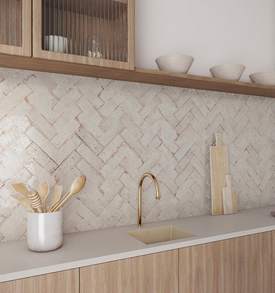 Modern kitchen with light wood cabinets, beige countertop, gold faucet, and herringbone-patterned backsplash tiles. Utensils are in a metal holder, with cutting boards and ceramic bowls on the counter and upper shelves.