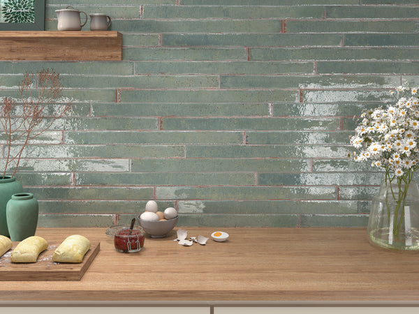 A kitchen countertop with dough, eggs, a jar of jam, and broken eggshells. Behind, glossy green tiles cover the wall, and shelves hold ceramic dishes and a vase. Fresh daisies sit in a jar on the right.