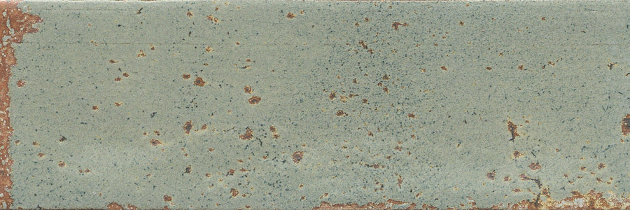 The Emerge 3x8 Porcelain Tile in Sprout features a rectangular shape with a weathered look, subtle surface texture, faded green tone, rust-like spots, and exposed brown accents along gently worn edges and across the tile.