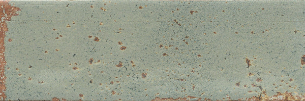 The Emerge 3x8 Porcelain Tile in Sprout features a rectangular shape with a weathered look, subtle surface texture, faded green tone, rust-like spots, and exposed brown accents along gently worn edges and across the tile.
