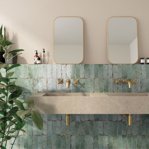 A modern bathroom features a double stone sink with gold fixtures, two rectangular mirrors, green mosaic tile backsplash, bottles of soap, and a large leafy plant in the corner.