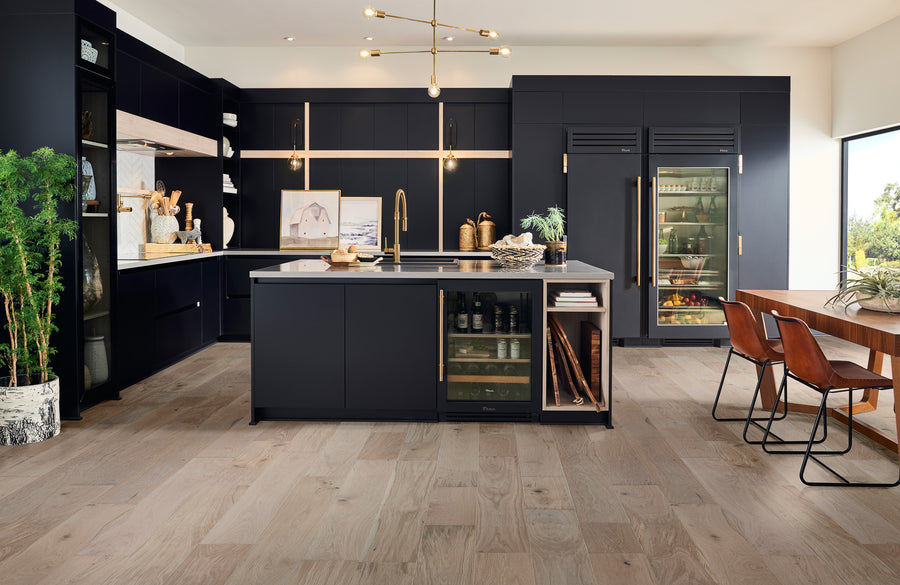 Modern kitchen with black cabinets, gold accents, and a large island. There are built-in refrigerators, open shelves with decor, a potted plant, and a wooden dining table with brown chairs by a large window.