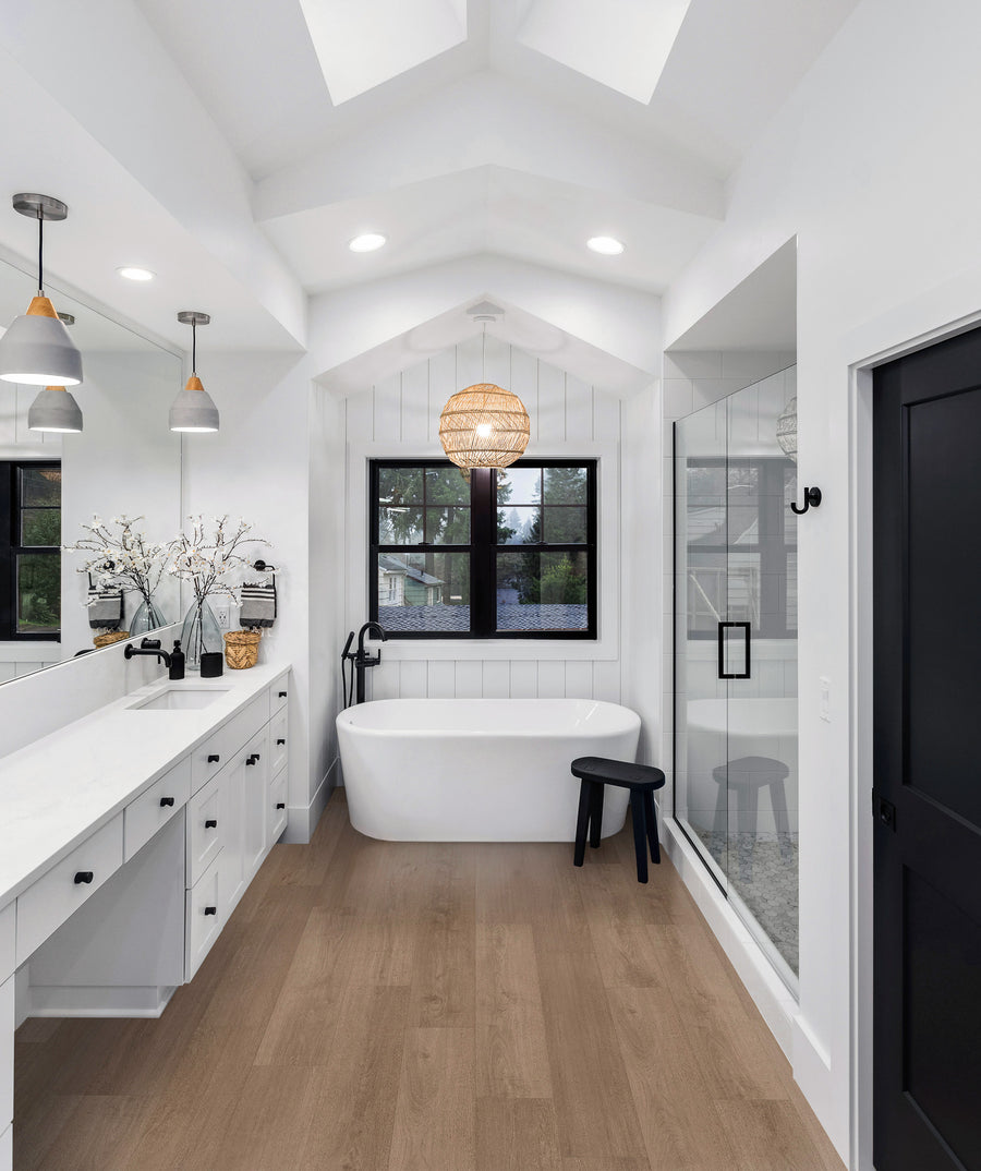 Modern bathroom with a freestanding white tub under a window, glass shower on the right, double sinks with a large mirror on the left, white walls, wood floor, black accents, and pendant lights.
