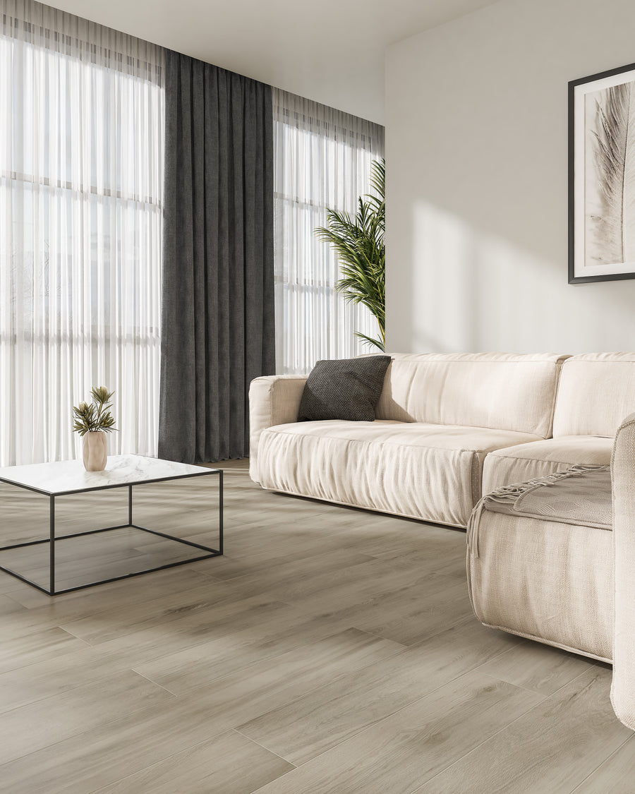 A modern living room with light wood flooring, a beige sofa, a black and white coffee table, a potted plant, dark curtains, and large windows with sheer white drapes letting in natural light.