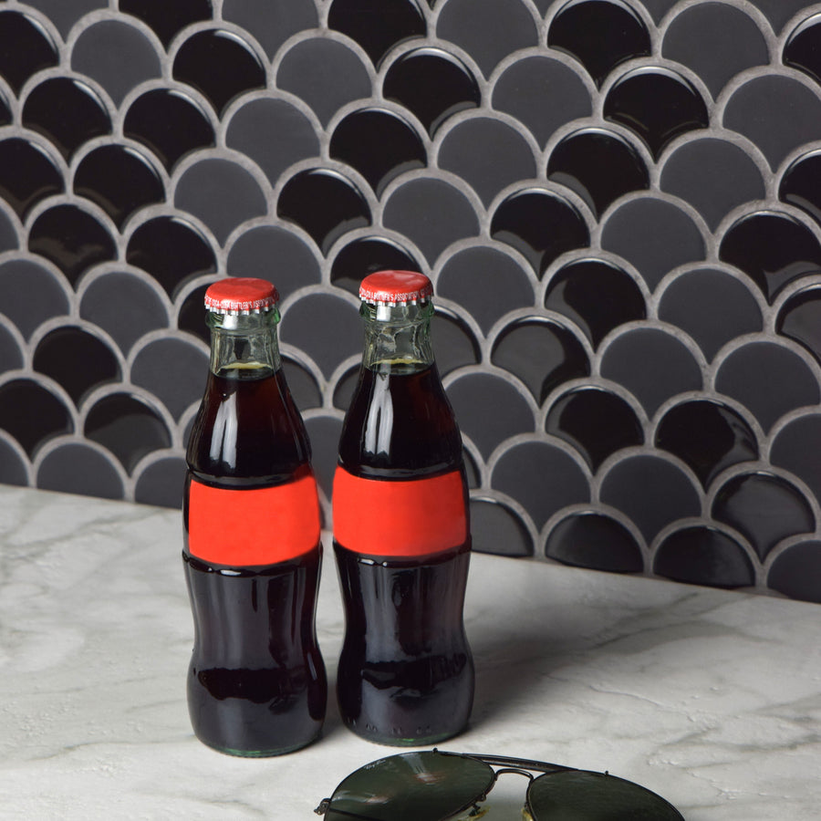 Two glass soda bottles with red labels sit on a marble surface in front of a dark scallop-patterned tile wall. A pair of sunglasses is placed nearby on the table.
