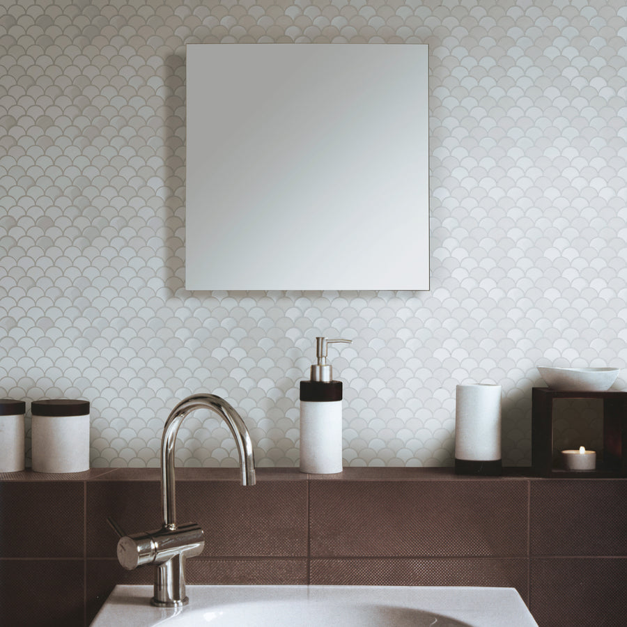 A modern bathroom with a round sink, a silver faucet, soap dispenser, candle, and containers on a brown counter. The wall features a subtle, light-colored fish scale tile pattern and a square mirror above the sink.