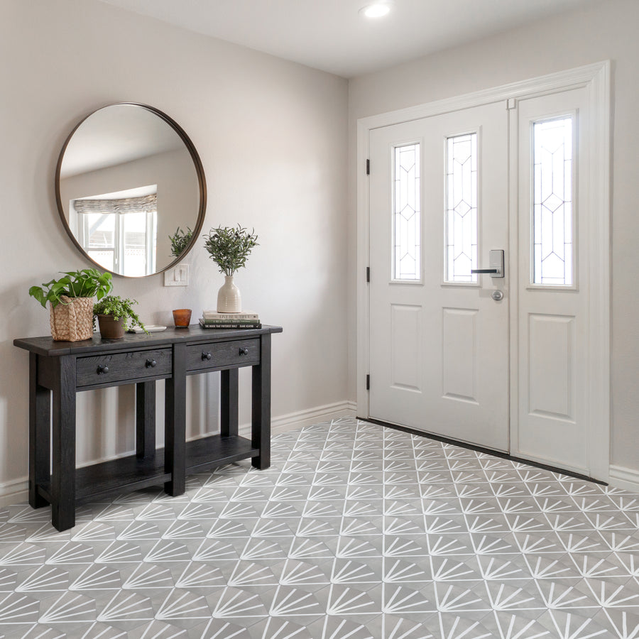 A bright entryway with a white front door, a round mirror, a dark wood console table, potted plants, books, and decorative items. The floor has a gray and white geometric patterned tile.