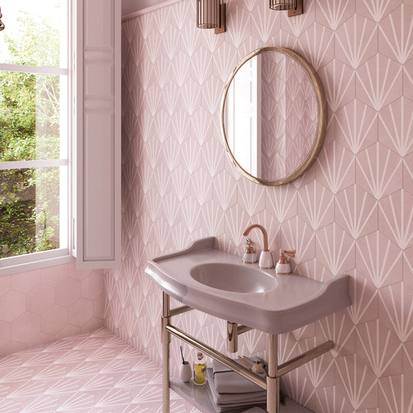 A stylish bathroom with geometric pink tiles, a gray sink, gold mirror, soap dispensers, neatly folded towels, and sunlight coming through an open window.
