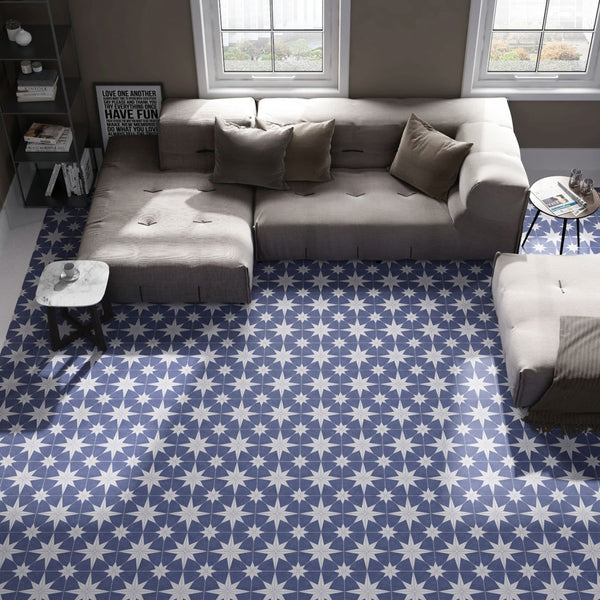 A top view of a modern living room with a beige L-shaped sofa, patterned blue and white star tile flooring, a small round table, a marble side table, a shelf with books, and two large windows letting in natural light.