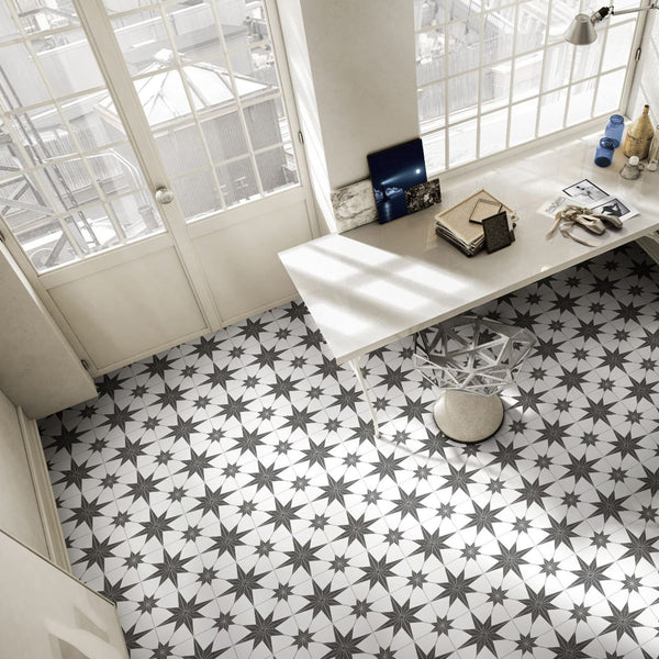 Bright room with large windows, a modern desk with books and decor, a metal stool, and patterned floor tiles featuring black and white star designs. Natural light fills the space, creating a clean, airy atmosphere.