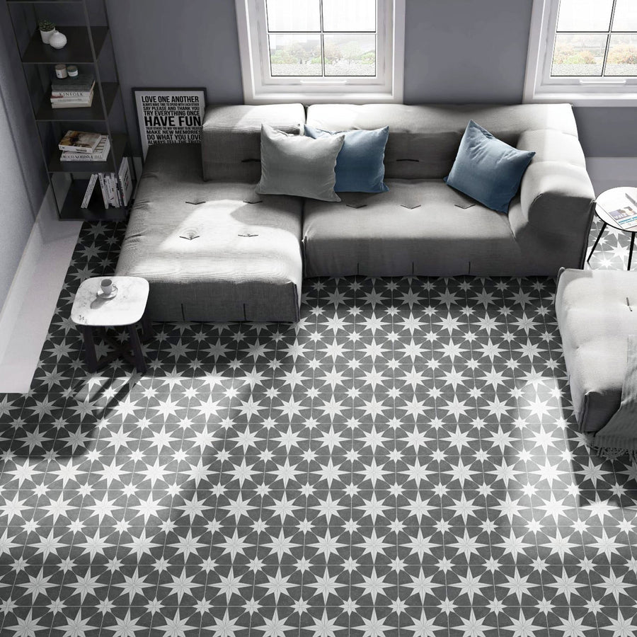 A modern living room with a gray sectional sofa, blue and gray cushions, a small round table, and large windows. The floor has a bold black and white starburst patterned tile.