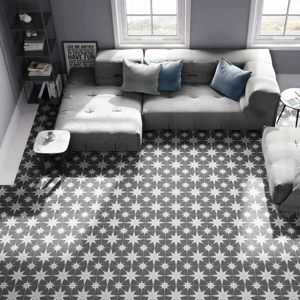 A modern living room with a gray sectional sofa, blue and gray cushions, a small round table, and large windows. The floor has a bold black and white starburst patterned tile.