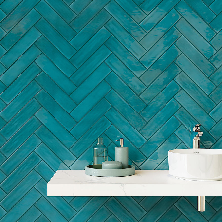 A white floating bathroom sink with a round basin and a chrome faucet sits against a wall covered in glossy teal herringbone tiles. On the sink, there are green bathroom accessories and a tray.