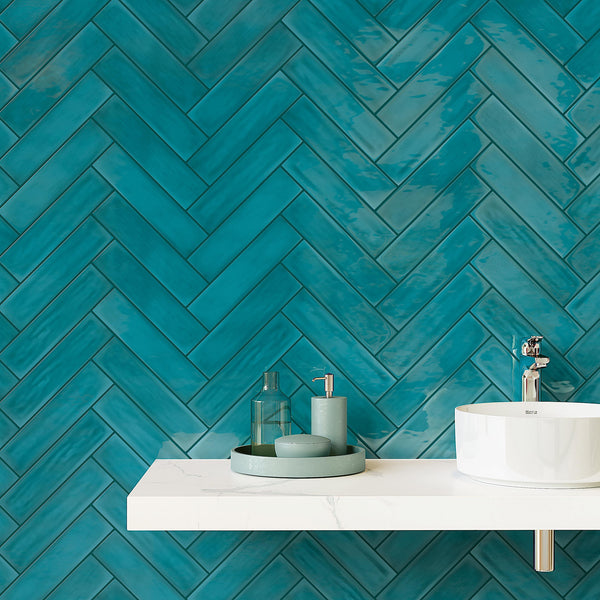A white floating bathroom sink with a round basin and a chrome faucet sits against a wall covered in glossy teal herringbone tiles. On the sink, there are green bathroom accessories and a tray.