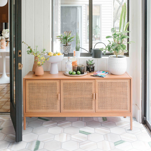 A wooden sideboard with woven cane doors is topped with lemons, a potted plant, a cutting board with limes, a bottle, glassware, and a watering can. Sunlight streams in through large windows onto a patterned tile floor.