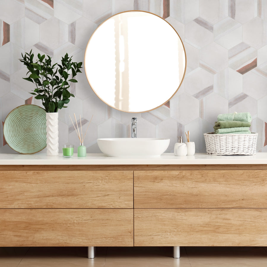 Modern bathroom vanity with light wood drawers, a white countertop, round mirror, potted plant, decorative plate, toiletries, and a white basket holding folded green towels, against a geometric tiled wall.