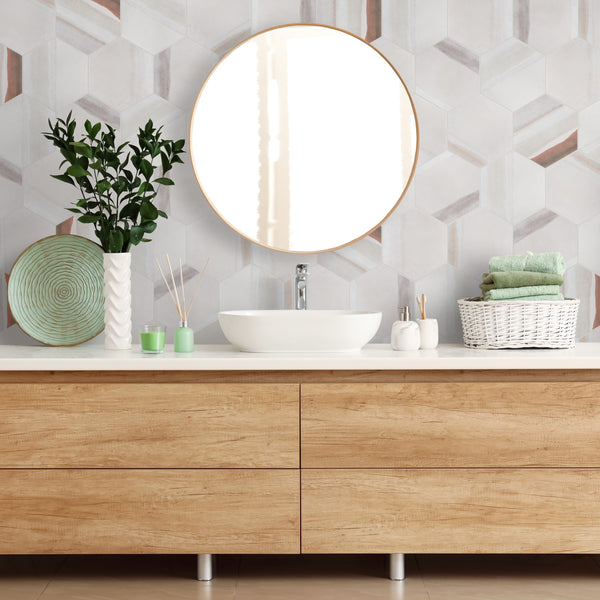 Modern bathroom vanity with light wood drawers, a white countertop, round mirror, potted plant, decorative plate, toiletries, and a white basket holding folded green towels, against a geometric tiled wall.