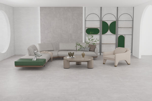 A modern living room with light gray floors and walls, a light gray sectional sofa with green ottoman, a beige armchair, a round coffee table, decorative plants, and minimalist shelving with green accents.