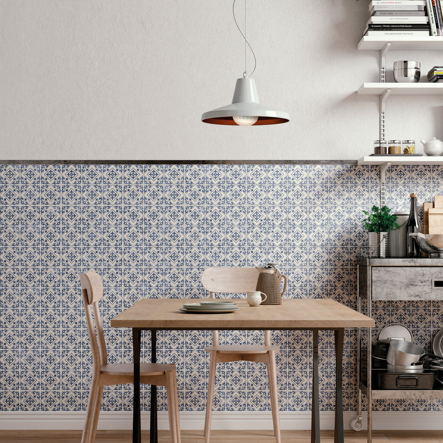 A small dining area with a wooden table and two chairs, set against a wall with blue and white patterned tiles. Above hangs a modern pendant light, and nearby are shelves with kitchen items and a metal cart with plants.