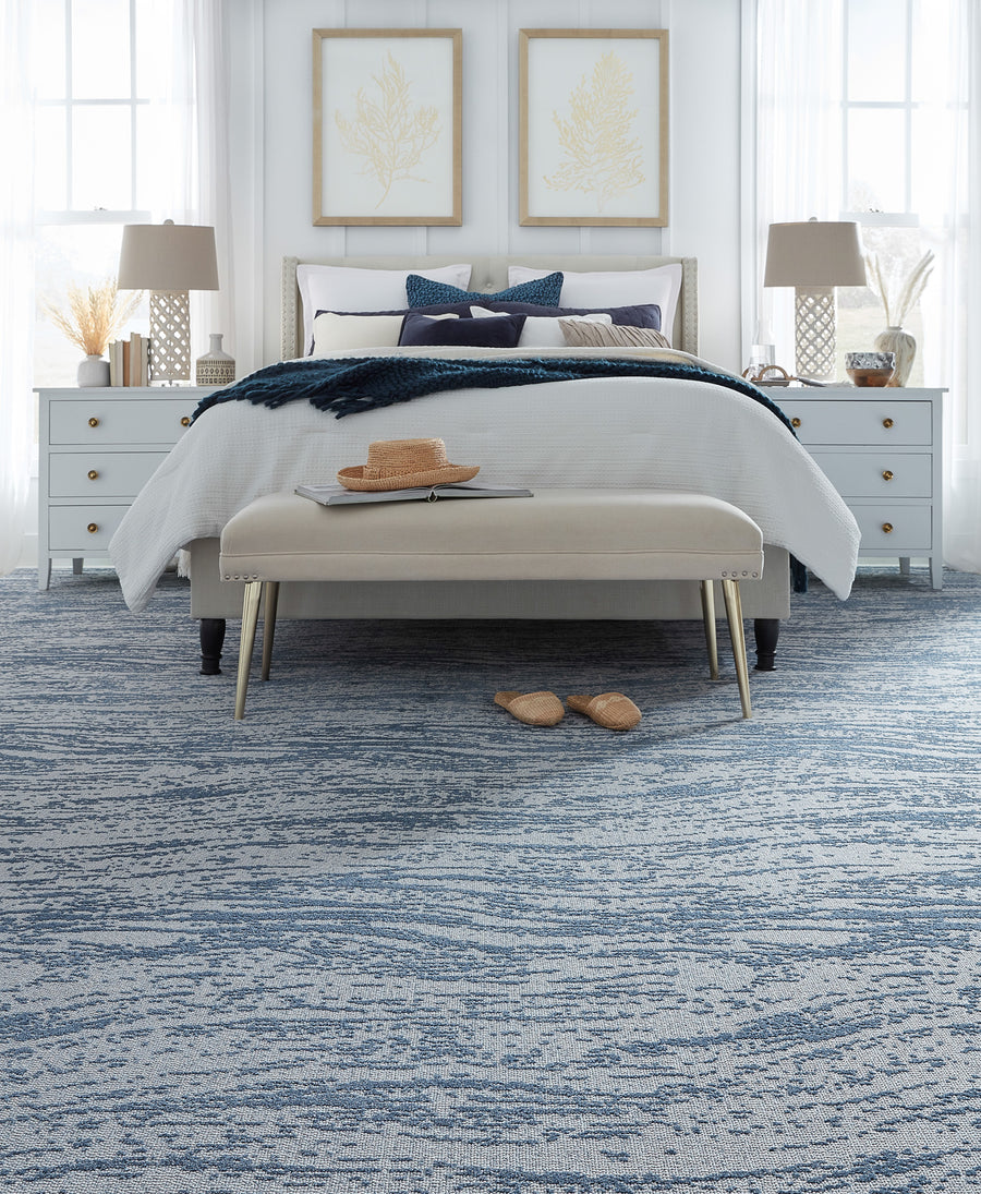 A neatly made bed with white and blue bedding stands between two white nightstands with lamps. A beige bench with a hat and tray sits at the foot, and slippers rest on a blue patterned carpet. Light fills the bright room.