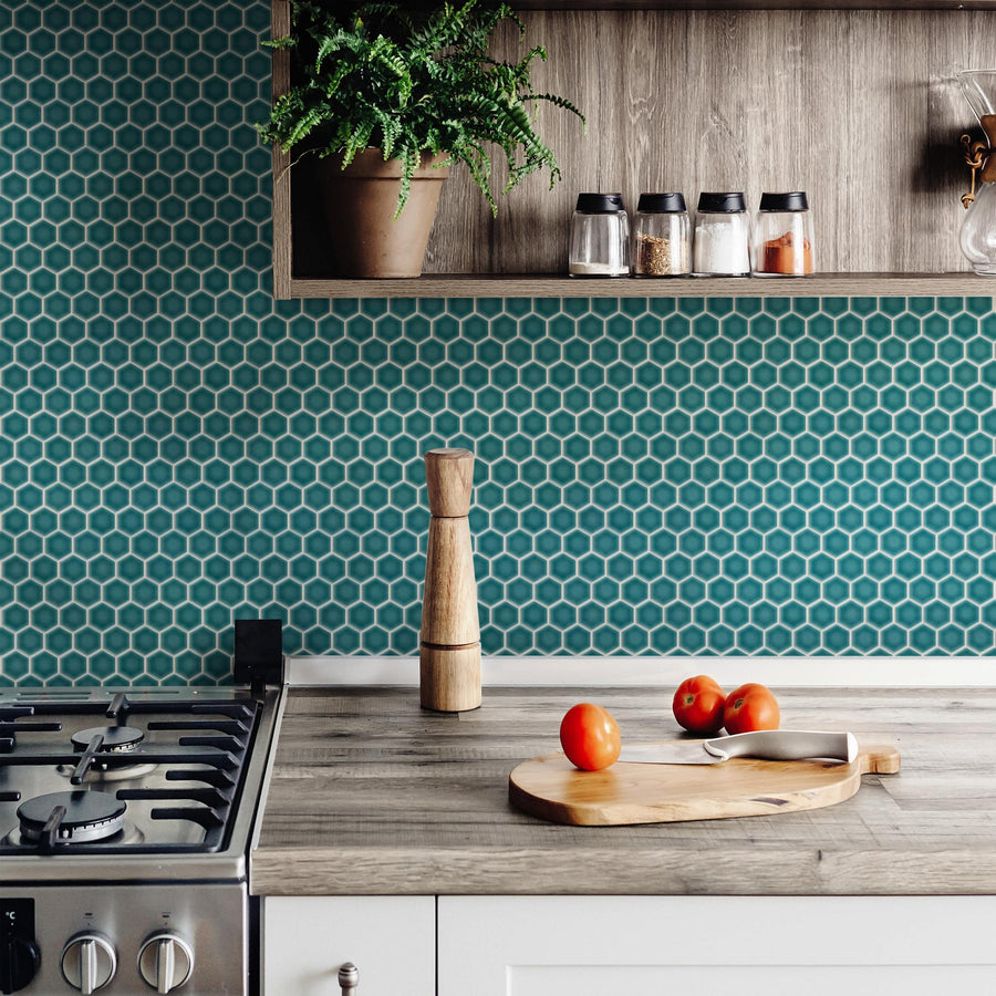 A modern kitchen with a teal hexagon tile backsplash, a wooden pepper mill, a cutting board with three tomatoes, a stovetop, and a shelf holding jars and a potted plant.