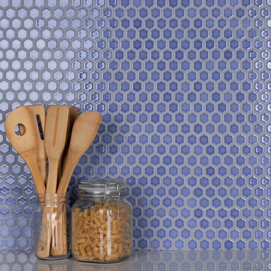 Wooden kitchen utensils in a glass jar and a closed glass jar filled with pasta sit on a counter in front of a blue hexagon tile backsplash.