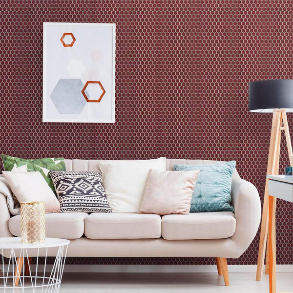 A modern living room with a beige sofa decorated with patterned cushions, a tripod lamp, a white side table, and a geometric art piece on a wall covered with small hexagonal maroon tiles.